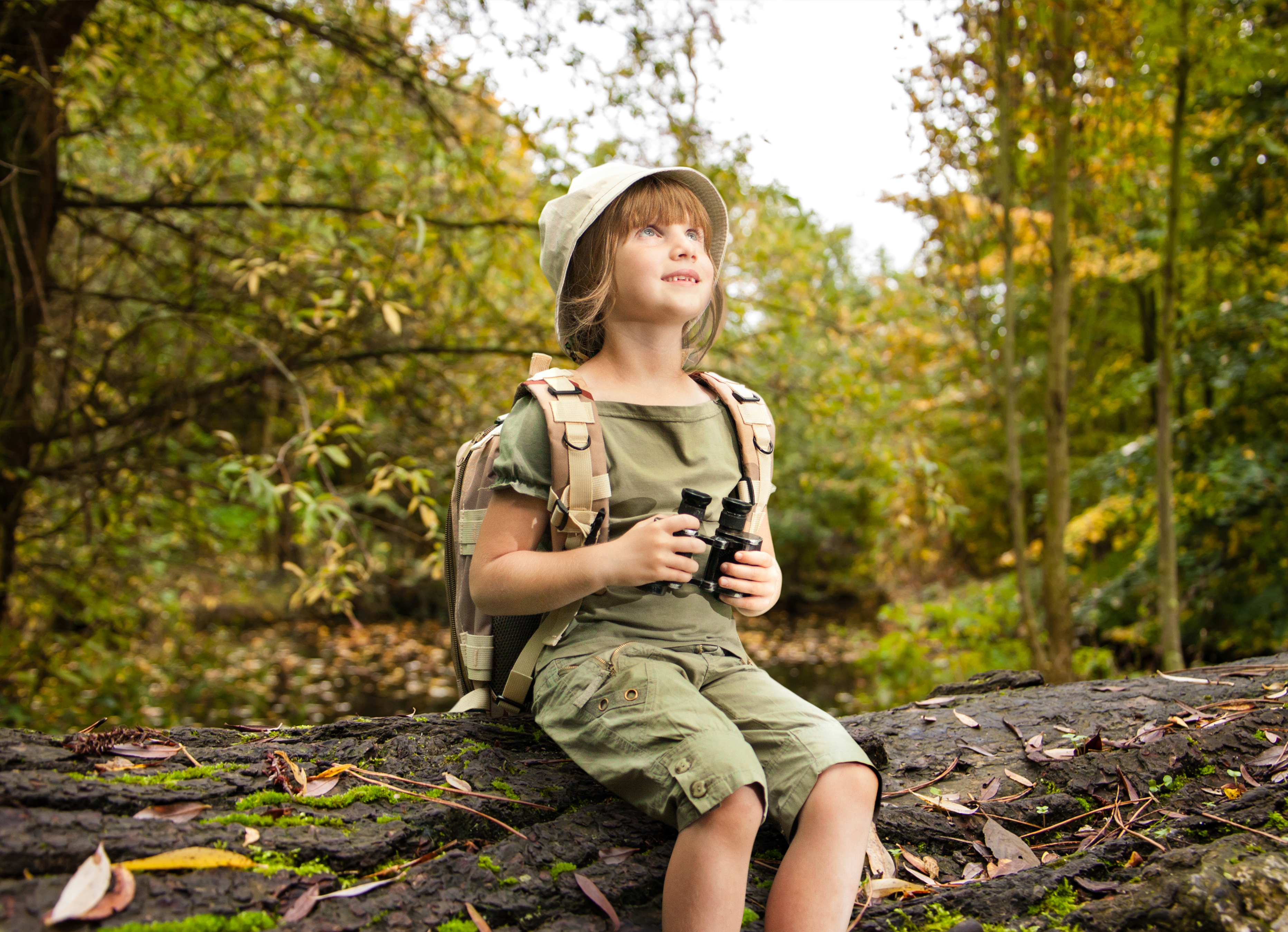 March 12 Special Events - Girl Scout Day. Girl sitting on a log in the forest holding a pair of binoculars and looking optimistically upwards. 